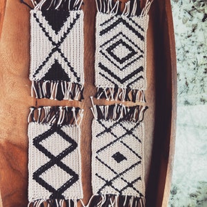 May include: Four black and white woven coasters with geometric patterns. The coasters have fringe around the edges.