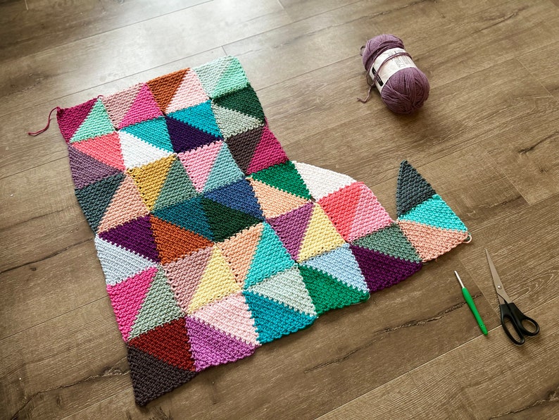 May include: A colorful crocheted blanket with a geometric pattern of triangles in shades of pink, purple, teal, green, and brown. The blanket is partially finished and laid out on a wooden floor. There is a ball of yarn and a pair of scissors nearby.