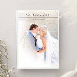 May include: A white wedding guide book with the title "WEDDING GUIDE" in a serif font. The cover features a photo of a bride and groom embracing. The book is on a light gray surface with white flowers and a white ribbon.