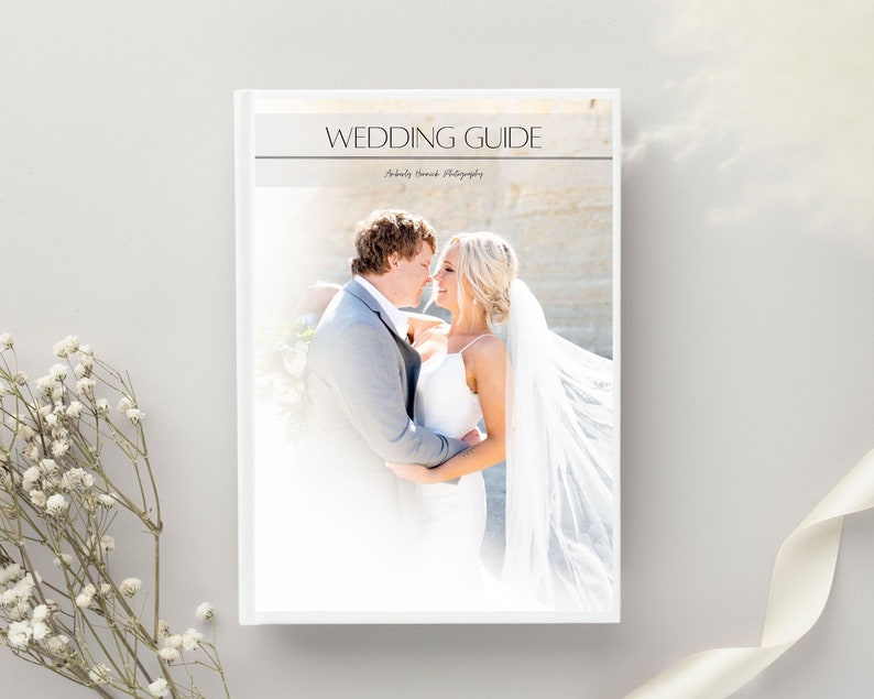 May include: A white wedding guide book with the title "WEDDING GUIDE" in a serif font. The cover features a photo of a bride and groom embracing. The book is on a light gray surface with white flowers and a white ribbon.
