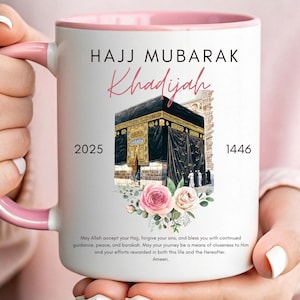 May include: White ceramic mug with a pink rim and handle. The mug features a graphic of the Kaaba, a floral design, and the text "Hajj Mubarak Khadijah 2025 1446". The text below the graphic reads "May Allah accept your Hajj, forgive your sins, and bless you with continued guidance, peace, and barakah. May your journey be a means of closeness to Him and your efforts rewarded in both this life and the Hereafter. Ameen."