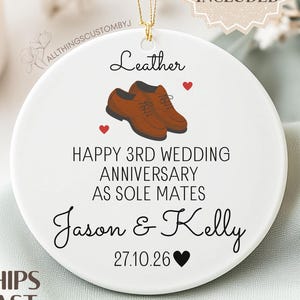 May include: A white ceramic ornament with a gold string for hanging. The ornament features a pair of brown leather shoes, hearts, and text that reads "Happy 3rd Wedding Anniversary as Sole Mates" and names. A gift box is included.