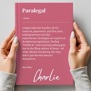 May include: A pink book with the word "Paralegal" in white text on the cover. The definition of the word is written in black text. The book also has the word "Charlie" written in black cursive text on the bottom of the cover.