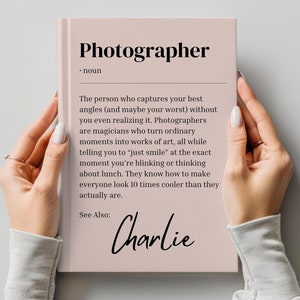 May include: A pink book with the word "Photographer" in black text on the cover. The definition of the word is written in black text below. The text "See Also: Charlie" is written in black cursive text at the bottom of the book.