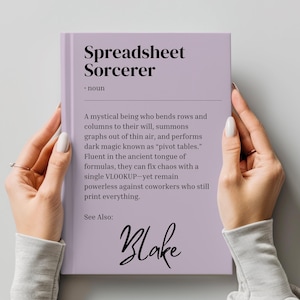 Op de afbeelding: Een paars boek met de titel "Spreadsheet Sorcerer" en de tekst "Een mystiek wezen dat rijen en kolommen naar zijn wil buigt, grafieken uit het niets oproept en donkere magie uitvoert die bekend staat als "pivot-tabellen."  Vloeiend in de oude taal van formules, kan het chaos oplossen met een enkele VLOOKUP-maar blijft machteloos tegen collega's die nog steeds alles printen." Het boek heeft ook de tekst "Zie ook: Blake" onderaan.