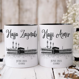 May include: Two white ceramic mugs with black rims and handles. The mugs feature a black and white illustration of the Kaaba in Mecca, Saudi Arabia. The text "Hajja Zaynab" and "Hajji Amir" are printed on the mugs, along with the date "June 2025 (1446)".
