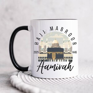 以下が含まれることがあります： 黒い取っ手付きの白いセラミック製のコーヒーカップ。カップには、サウジアラビアのメッカにあるカアバのグラフィックと、「Hajj Mabroor 2025 1446 Aamivah」というテキストが描かれています。