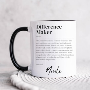 Può includere: Una tazza in ceramica bianca con manico e bordo neri. La tazza presenta una definizione in bianco e nero della parola "Difference Maker" con il nome "Nicole" scritto in corsivo in basso.
