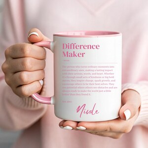 Peut inclure: Une tasse en céramique blanche avec un bord et une poignée roses. La tasse présente une définition rose du mot "Difference Maker" et le nom "Nicole" en bas.