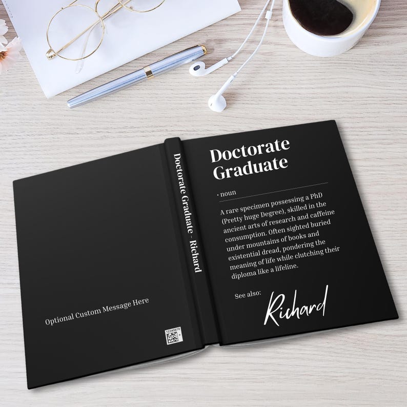 以下が含まれることがあります： 「Doctorate Graduate」というテキストと、その用語の定義が書かれた黒い本。ページの下部に「Richard」という名前が筆記体で書かれている。