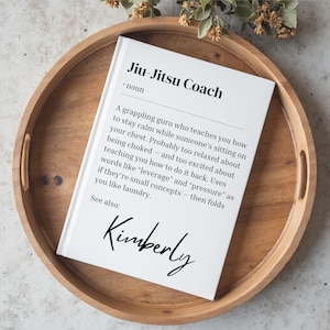 Puede incluir: Un cuaderno blanco con el texto "Jiu-Jitsu Coach" y una definición, sobre una bandeja de madera. La definición describe a un entrenador de Jiu-Jitsu. El nombre "Kimberly" está escrito en la parte inferior del cuaderno.