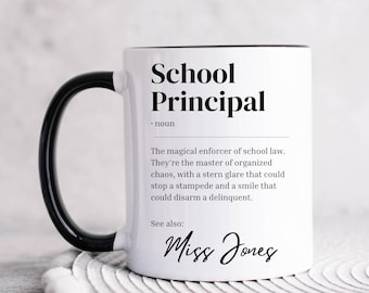 Puede incluir: Taza de cerámica blanca con asa y borde negros. La taza tiene un diseño de texto negro que dice "School Principal" y una definición de la palabra. El texto "See also:" va seguido del nombre "Miss Jones" en cursiva.