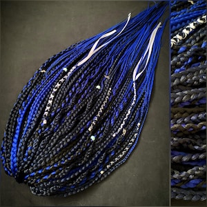 May include: A collection of blue and black synthetic dreadlocks, decorated with silver chains, beads, and white ribbons. The dreadlocks are displayed on a dark surface, highlighting the detailed braiding and colour gradient.