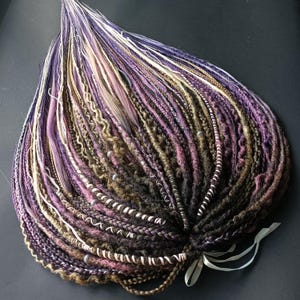 May include: A collection of synthetic dreadlocks in purple, brown, and white hues. The dreadlocks vary in thickness and texture, with some featuring beads and wrapped details. A white ribbon is tied at the base of the dreadlocks.