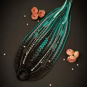 Puede incluir: Un conjunto de rastas negras, turquesas y grises, adornadas con cuentas, plumas y detalles plateados. Las rastas están dispuestas sobre una superficie negra, con rosas color melocotón y perlas dispersas.