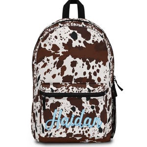 May include: A brown and white cow print backpack with a black handle and zipper. The name "Haidan" is embroidered in light blue on the front pocket. The backpack has a mesh side pocket.