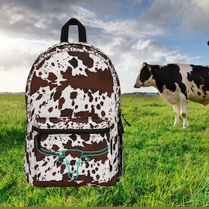 May include: A brown and white cow print backpack with a turquoise bull head design. The backpack is in front of a green grassy field with two black and white cows in the background.