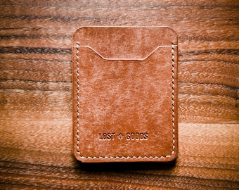 May include: A brown leather card holder with a stitched edge and the text "LOST GOODS" embossed on the front.
