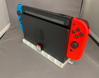 May include: A white stand with a Nintendo Switch console and a red Joy-Con controller. The console is in its dock and the Joy-Con is attached to the right side. The stand has a slot for a game cartridge.