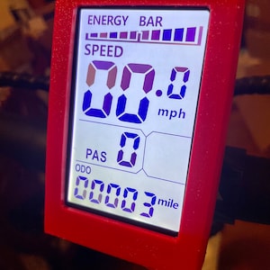 May include: A red digital display with a white background shows the speed of an electric bike. The display shows a speed of 0.0 mph and an odometer reading of 0.0003 miles. The display also shows a bar graph for energy level.
