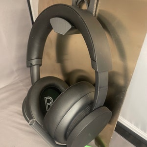 May include: Black over-ear headphones with a built-in microphone are suspended from a 3D-printed holder. The holder is attached to a metallic plate and features the Xbox logo. The headphones have a padded headband and earcups.