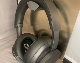 May include: Black over-ear headphones with a built-in microphone are suspended from a 3D-printed holder. The holder is attached to a metallic plate and features the Xbox logo. The headphones have a padded headband and earcups.