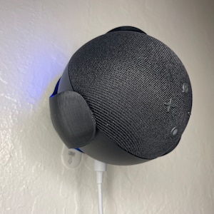 Op de afbeelding: Een zwarte slimme speaker met een stoffen rooster en een wit stroomsnoer. De speaker is gemonteerd aan een witte muur.