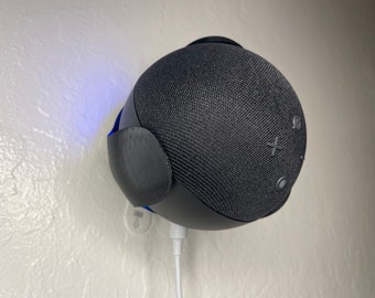 May include: A black smart speaker with a fabric grille and a white power cord. The speaker is mounted on a white wall.