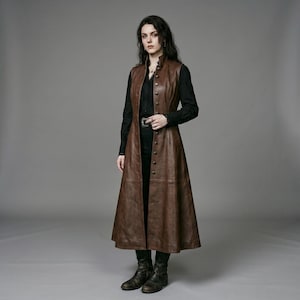 May include: A long, sleeveless brown leather vest featuring a high collar and a vertical row of metallic buttons down the front. The garment is worn over a black long-sleeved shirt and black trousers, creating a layered look with a fitted bodice.