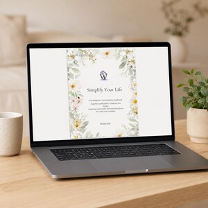 May include: A laptop screen displaying a digital workbook titled Simplify Your Life. The cover features a white background with a floral border of white flowers and green leaves, a purple logo, and the text Wilderwell at the bottom.