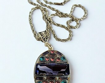 May include: Silver-toned necklace with an oval pendant. The pendant features a large, rectangular, dark purple stone and is surrounded by smaller, colorful gemstones in shades of pink, green, and purple. The chain is a twisted rope design.