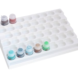 Peut inclure: Porte-pots de peinture en plastique blanc avec 48 fentes individuelles pour ranger de petits pots de peinture. Le porte-pots a un rebord surélevé sur le bord avant et un petit rebord sur le bord arrière. Il y a six pots de peinture dans le porte-pots, dont cinq sont bleus, verts et bruns. Le sixième pot est rouge.