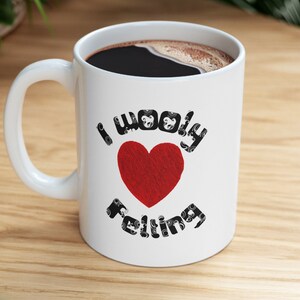 May include: White ceramic mug with a black handle. The mug has a red heart with the text "I woolly felting" in black letters around the heart.