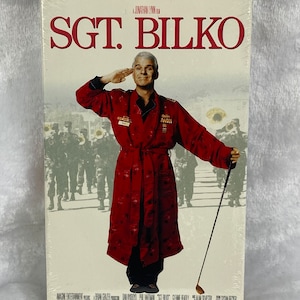 May include: A VHS tape cover featuring Steve Martin in a red robe, saluting, with the title "Sgt. Bilko" and the text "A Jonathan Lynn Film".