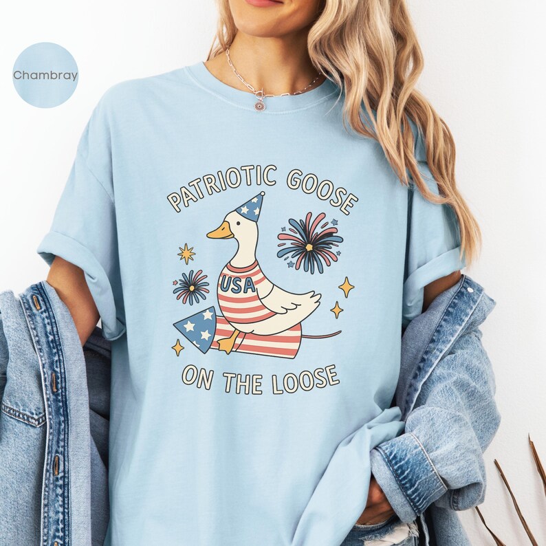 May include: Light blue t-shirt with a patriotic goose graphic. The design features a goose wearing a party hat and a USA-themed outfit, with fireworks and stars. Text reads "Patriotic Goose On The Loose."