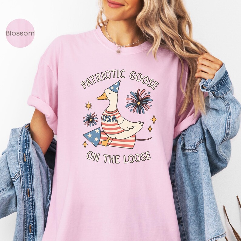 May include: A light pink t-shirt featuring a patriotic goose design. The goose wears a party hat and a USA-themed shirt, with fireworks and the text "PATRIOTIC GOOSE ON THE LOOSE". The shirt is a soft, comfortable material.