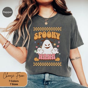 Può includere: T-shirt grigio scuro con un design a tema Halloween. Il design presenta la parola "Spooky" in arancione, un fantasma bianco, due fantasmi più piccoli e la parola "Nana" in rosa. La maglietta ha uno stile retrò e vintage.