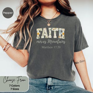 May include: Dark grey t-shirt featuring the text "FAITH moves Mountains" in a floral design. The text "Matthew 17:20" is below. Available in 7 colours and 7 sizes.