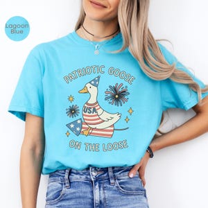May include: Lagoon blue t-shirt with a patriotic goose graphic. The design features a goose wearing a party hat and a USA-themed outfit, with fireworks and stars. Text reads "Patriotic Goose On The Loose."