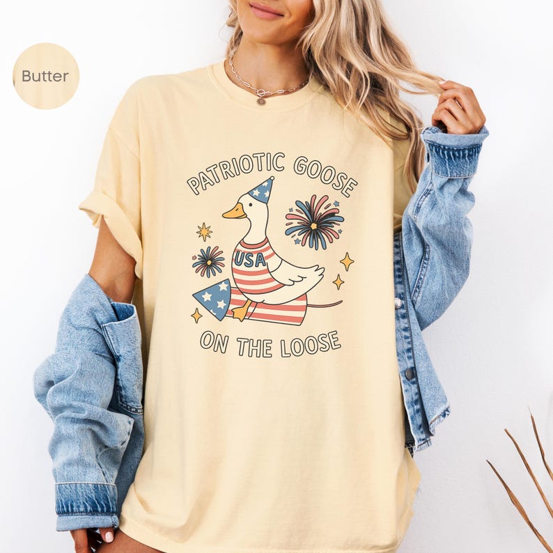 May include: Butter-colored t-shirt with a patriotic goose graphic. The goose wears a USA-themed outfit and a party hat, with fireworks and stars. Text reads "Patriotic Goose On The Loose."