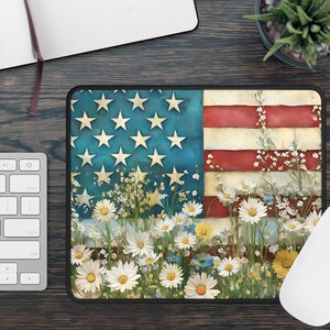 May include: Rectangular mousepad featuring a stylized American flag design. The flag's blue field with white stars and red and white stripes is adorned with a field of daisies and wildflowers. The mousepad is on a dark wooden surface.