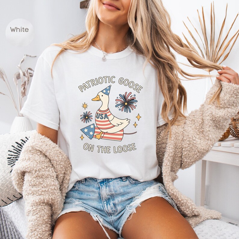 May include: White t-shirt featuring a patriotic goose design. The graphic includes a goose wearing a party hat and a USA-themed outfit, with the text "PATRIOTIC GOOSE ON THE LOOSE". The design incorporates fireworks and stars.