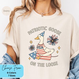 May include: Ivory-colored t-shirt featuring a patriotic goose design. The goose wears a party hat and a USA-themed outfit, with fireworks and stars. Text reads "Patriotic Goose On The Loose".