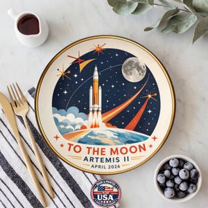 May include: A round ceramic plate with a space-themed design. The plate features a rocket launching towards the moon, with the text "TO THE MOON ARTEMIS II APRIL 2026". The plate has a gold rim and is surrounded by a white background.