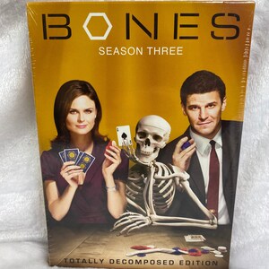 May include: A DVD box set for the TV show "Bones" season three. The box features a yellow background with the show's title in white and black. The image shows the two main characters, a woman and a man, sitting at a table with a skeleton. The text "Totally Decomposed Edition" is at the bottom of the box.