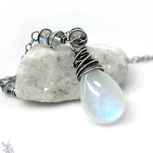 May include: A silver-toned necklace featuring a teardrop-shaped, iridescent moonstone pendant. The pendant is wire-wrapped and suspended from a delicate chain. Small, round moonstone beads accent the chain near the clasp. The necklace rests on a white stone.