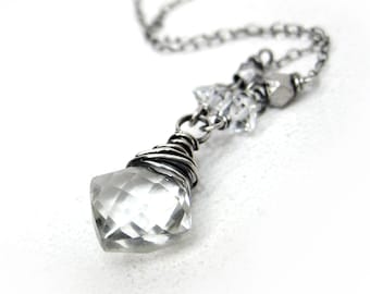 May include: A silver necklace with a clear, faceted crystal pendant. The crystal is wire-wrapped and hangs from a delicate chain. Additional clear crystals and silver beads adorn the chain. The necklace is set against a white backdrop.
