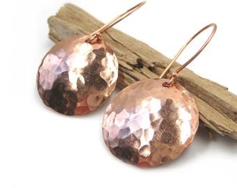 May include: A pair of copper-colored, hammered metal earrings. Each earring features a circular, textured pendant suspended from a simple ear wire. The earrings are displayed on a piece of driftwood against a white background. The earrings have a rustic, handmade aesthetic.
