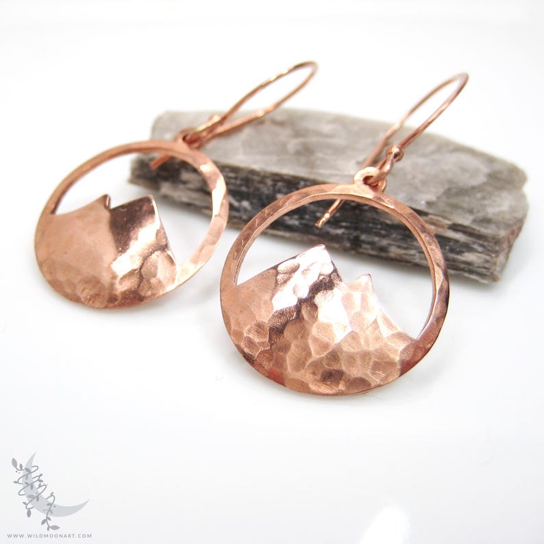 May include: A pair of hammered copper earrings with a mountain silhouette cut out. The earrings are round and have a hammered texture.