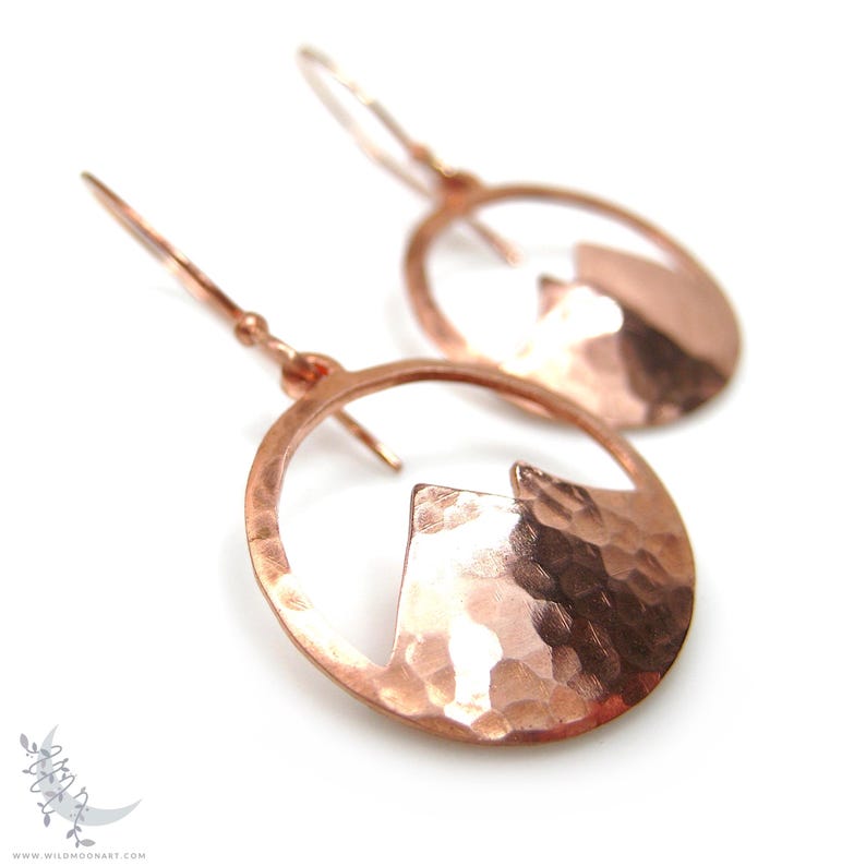 May include: A pair of copper earrings with a hammered finish. Each earring features a circular design with a cut-out mountain shape.